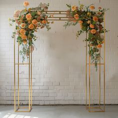 two gold metal plant stands with flowers on them against a white brick wall in front of a window