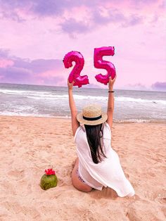 a woman sitting on the beach holding up pink numbers