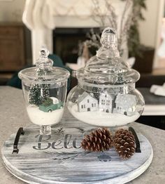 two glass jars filled with snow and pine cones on top of a tray next to a fireplace