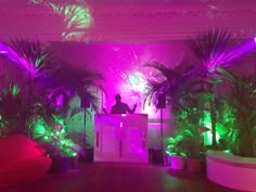 a man standing at a podium surrounded by potted plants and palm trees in a room with purple lighting