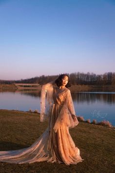 a woman is standing in front of a lake wearing a long dress with sheer sleeves