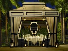 a gazebo with white table cloths and chairs under palm trees in the evening