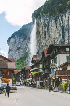 people are walking down the street in front of some buildings with a waterfall coming out of it