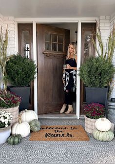 a woman standing in front of a door with potted plants and pumpkins on the doorstep