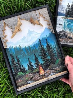 someone is holding up two paintings on the grass with trees and mountains in the background