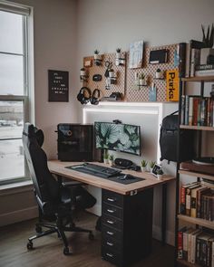 a desk with a computer on it in front of a window and bookshelf