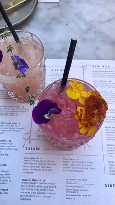 two cocktails with flowers in them on a table next to a drink glass and menu