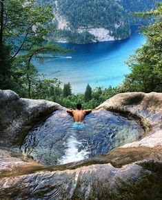 a man is swimming in a lake surrounded by mountains and trees, with his back turned to the camera