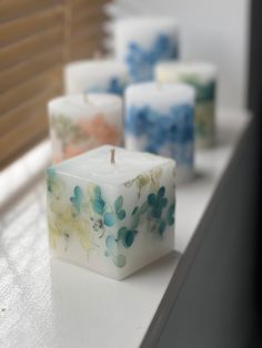 four candles are sitting on a window sill with flowers painted on them and one candle is in the shape of a cube