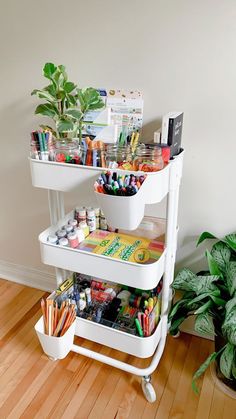 three tiered white cart with art supplies on it next to a potted plant