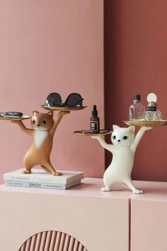 two cats holding trays on top of a pink shelf in front of a wall