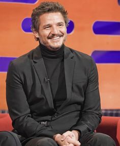 a smiling man in a black suit sitting on a red chair