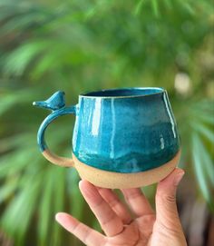 a hand holding a blue ceramic mug with a bird on it's handle, in front of a green plant