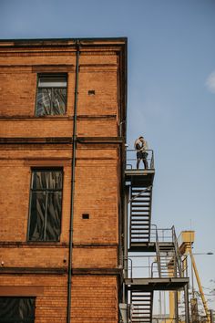 a man standing on top of a metal staircase next to a tall brick building with windows