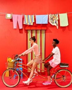 two people riding on a bike in front of a red wall with clothes hanging above it