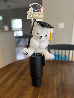 a white teddy bear wearing a graduation cap and gown sitting on top of a black cup