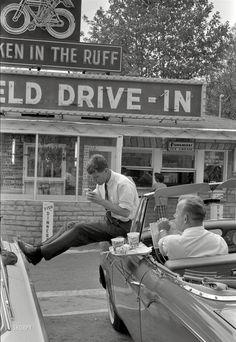 two men sitting on the hood of a car in front of an old drive - in