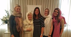 four women are standing together in front of a window and smiling at the camera, with one woman wearing a headscarf