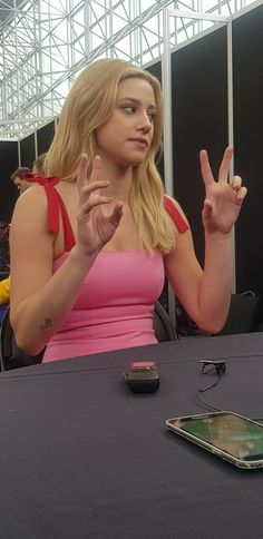 a woman sitting at a table in front of a cell phone and making the peace sign