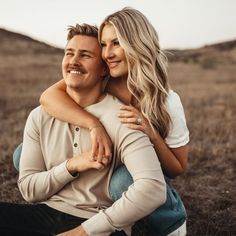 a man and woman are sitting on the ground in an open field, hugging each other