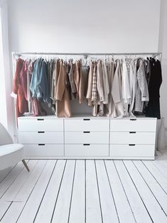 an organized closet with white drawers and clothes hanging on the rail, next to a chair