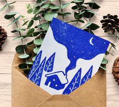 an envelope with a blue and white paper cut out of it sitting next to pine cones