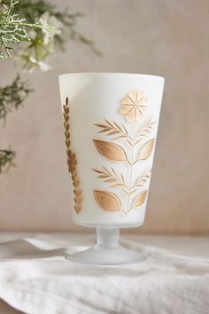 a white glass with gold leaf designs on it sitting next to a plant in a vase