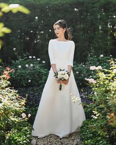 a woman in a white wedding dress standing in front of some bushes and flowers with her hands on her hips