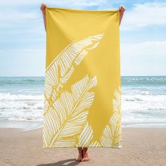 a person standing on the beach holding up a yellow towel with white palm leaf designs