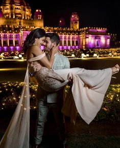 a man holding a woman in front of a building with lights all around it at night