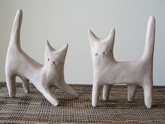 two white ceramic cats sitting next to each other on a tablecloth covered floor with a wall in the background