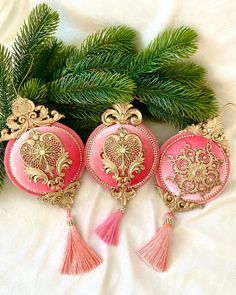 three pink ornaments with tassels are on a white surface next to pine branches