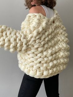 a woman in black pants and a white sweater is holding up a large chunky knitted blanket