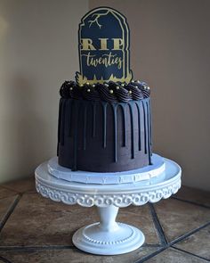 a black and white cake with gold lettering on top is sitting on a pedestal in front of a wall