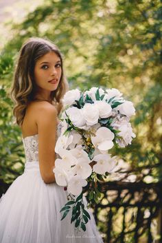 a woman in a wedding dress holding a bouquet of white flowers and greenery on her shoulder