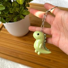 a hand holding a green plastic dinosaur keychain next to a potted plant