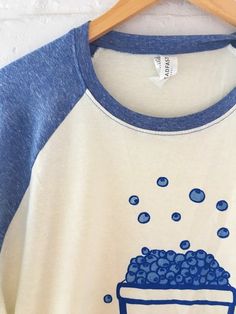 "Hand Printed and Hand Drawn! This is a raglan baseball tee printed with a hand drawn basket of blueberries. The shirt shown here is natural with triblend navy blue sleeves. // PROCESS: All of our items are individually hand printed by either me or my dad, which can lead to slight variations in placement. The ink is heat cured and will not fade over time. Since our items are printed to order, there is a 1-2 week processing time before shipping. If you absolutely need an item shipped quicker than Blue Raglan Sleeve T-shirt For Summer, Blue Relaxed Fit Top With Hand Printed Details, Blue Relaxed Fit Hand Printed Top, Blue Hand Printed Top With Relaxed Fit, Casual Blue Hand Printed Top, Cotton Raglan Sleeve T-shirt With Screen Print, Basket Of Blueberries, Blueberry Shirt, Gardening Gift