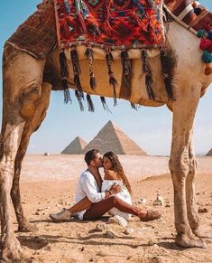 a man and woman sitting in front of a camel with pyramids in the background