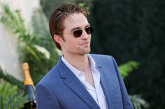 a man in a blue suit and sunglasses standing next to a bottle of wine with palm trees behind him