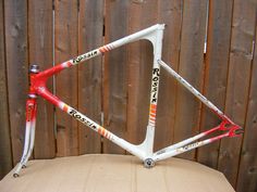 a white and red bike frame sitting on top of a cardboard box next to a wooden fence