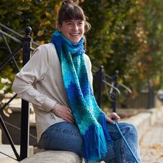 a woman sitting on a bench wearing a blue and green knitted scarf with tassels