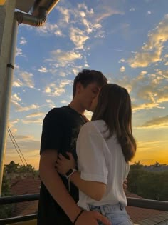 a young man and woman kissing on the balcony at sunset, with clouds in the background