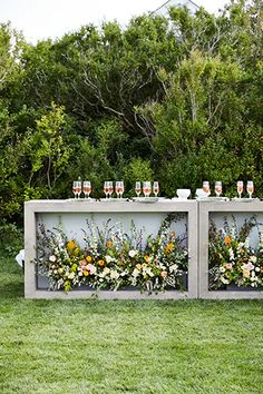 an outdoor bar with flowers and wine glasses on it's side, surrounded by greenery