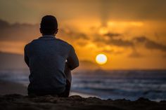 a man sitting on top of a sandy beach next to the ocean at sunset time