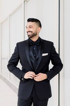 a man in a black tuxedo smiles while standing next to a white wall