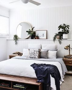 a bed with white sheets and pillows in a bedroom next to a mirror, plant on the wall