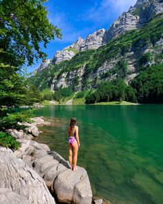 a woman standing on top of a rock next to a body of water with mountains in the background