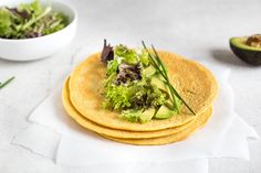 three tortillas with lettuce and avocado on top next to a bowl of salad