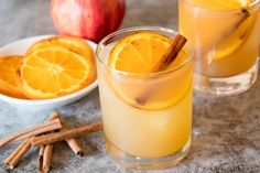 two glasses filled with orange juice and cinnamon sticks on a table next to an apple