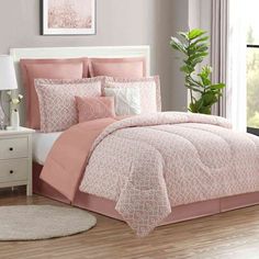 a bed with pink comforter and pillows in a room next to a plant on the floor